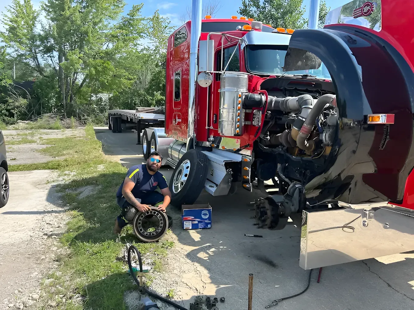 Man working on a red semi-truck with the hood open outdoors. The man is crouching, and a wheel is on the ground.