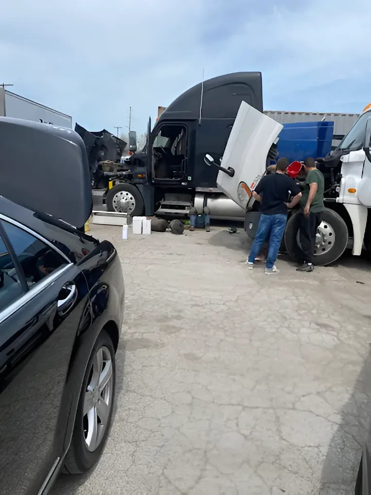 Men working on a black semi-truck with its hood open in an outdoor lot. A black car with its hood up is in the foreground.