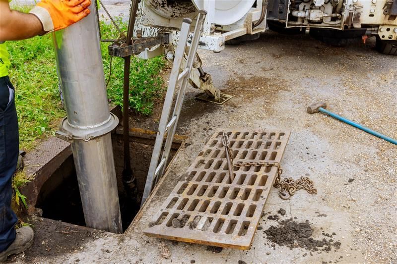 Worker in orange glove lowers pipe into open sewer. Metal grate and equipment visible.