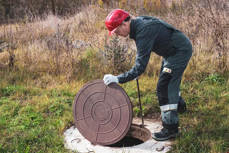 Worker in red hardhat opening a manhole cover in a grassy outdoor area.