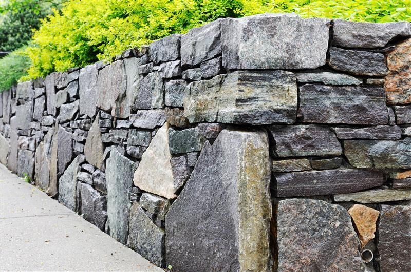 Stone retaining wall with gray and brown rocks, sidewalk, and green foliage.