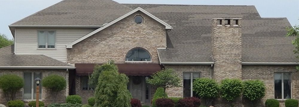 A large brick house with a gray roof is surrounded by trees and bushes.