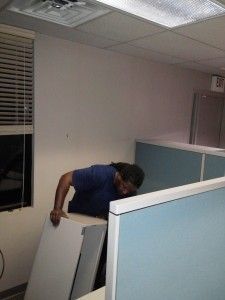 A man is holding a piece of wood in an office cubicle.