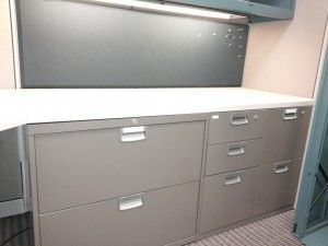 A cubicle with a desk , filing cabinets and drawers.