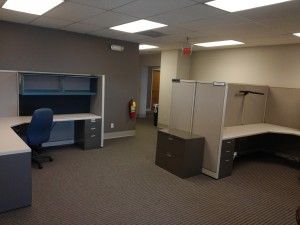 A cubicle in an office with two desks and a chair.