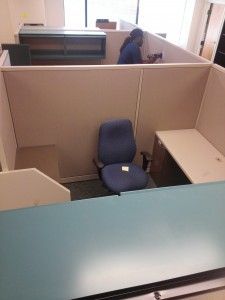 A woman is standing in a cubicle with a blue chair.