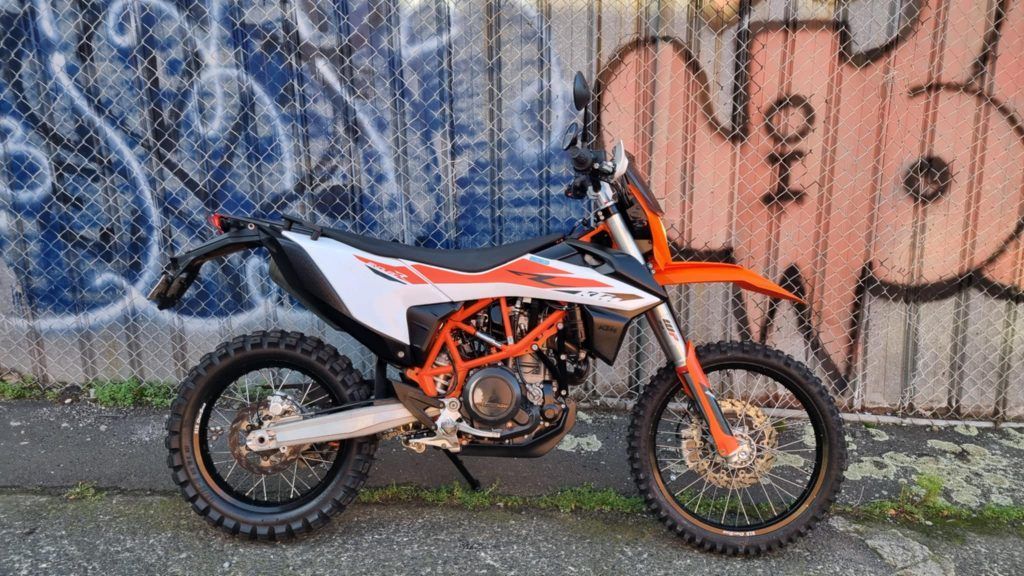 A white and orange KTM dual-sport motorcycle parked on an asphalt path in front of a chain-link fence with graffiti.