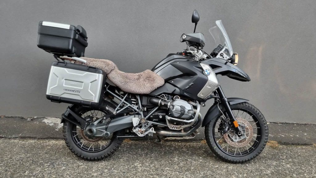 A black BMW adventure motorcycle with silver side panniers, a black top case, and a sheepskin seat cover parked outside.
