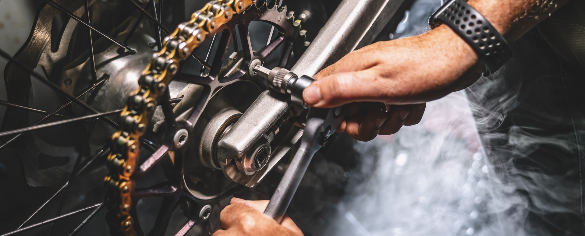 Close-up of hands using a wrench and socket to adjust the chain tensioner on a motorcycle rear wheel.
