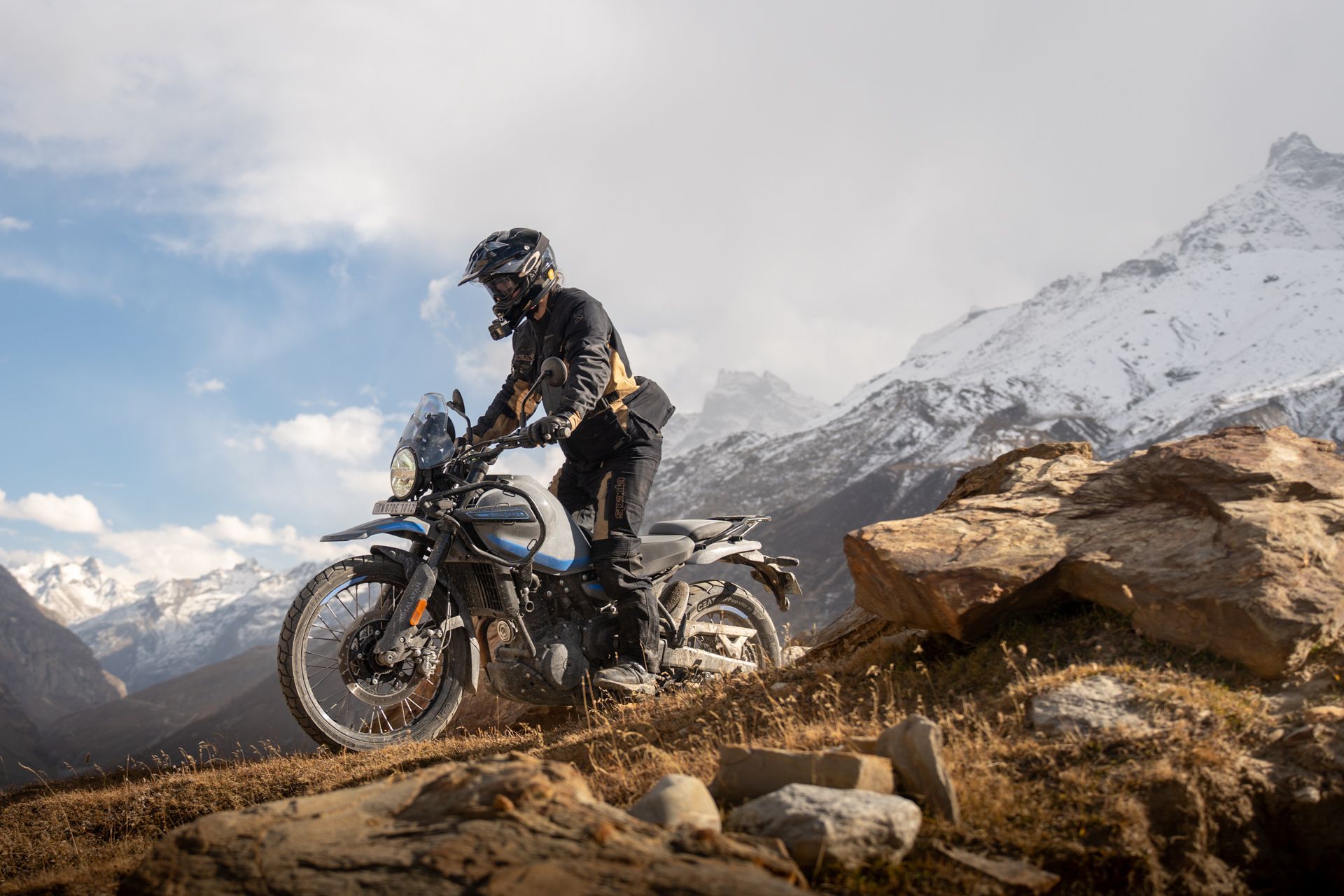 A motorcyclist rides a light-blue adventure bike across a rocky, grassy mountain slope with snow-capped peaks behind.