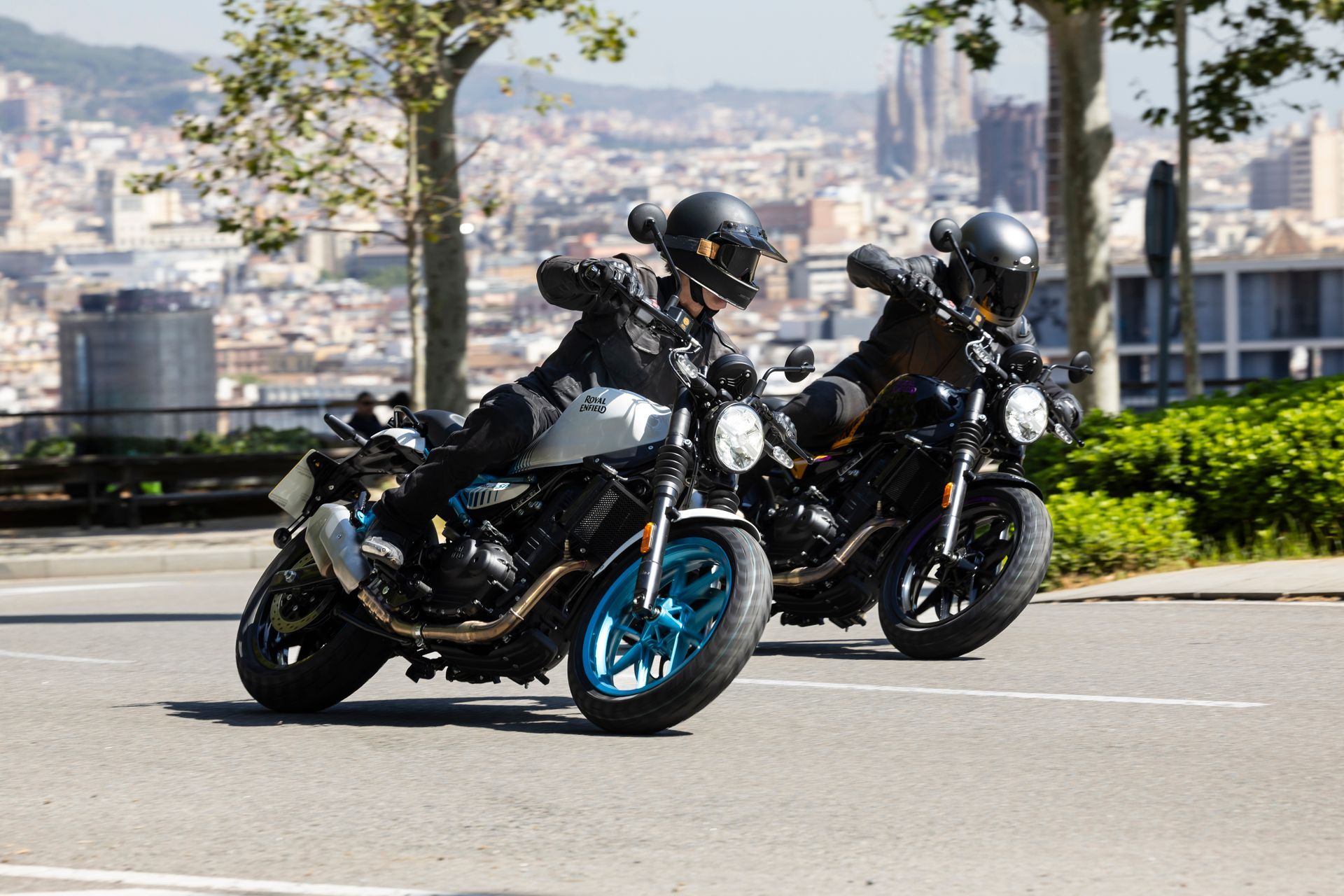 Two motorcyclists in helmets ride side-by-side on an urban road with a city skyline in the background.