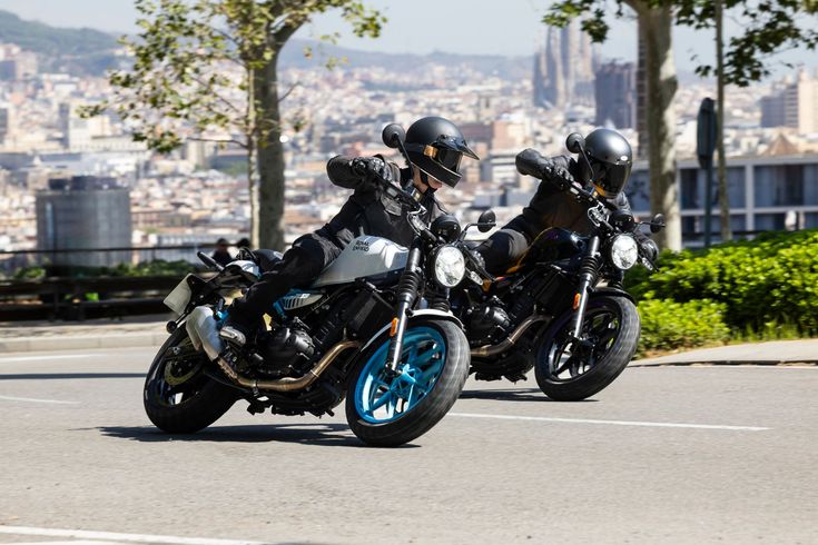 Two motorcyclists in helmets ride side-by-side on an urban road with a city skyline in the background.