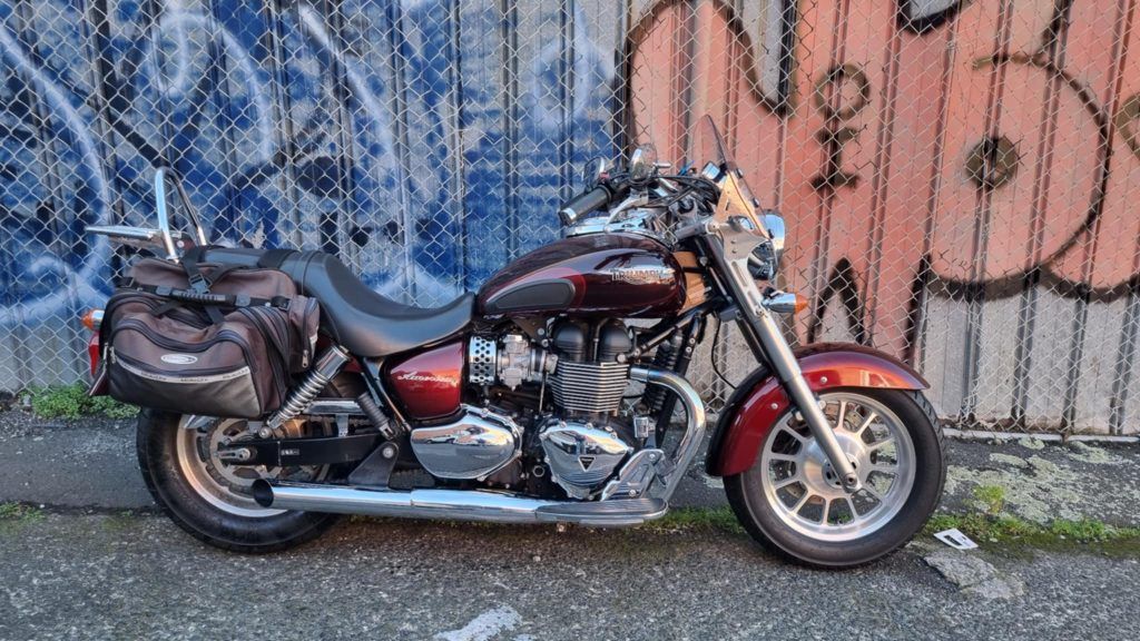 A maroon Triumph cruiser motorcycle parked on a paved street in front of a graffiti-covered chain-link fence.