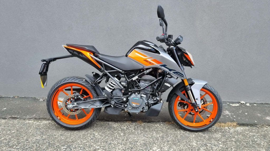 A silver and orange KTM Duke motorcycle parked against a grey wall on an asphalt surface.