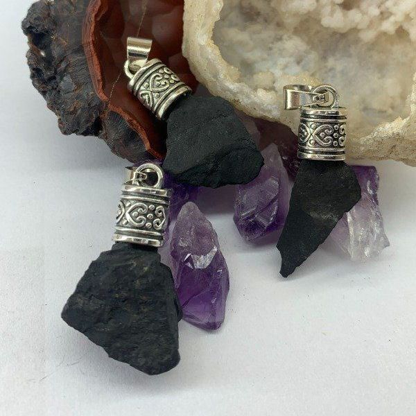 Un colgante de piedra preciosa negra en forma de lágrima con asa de plata, rodeado de cristales y rocas de amatista.