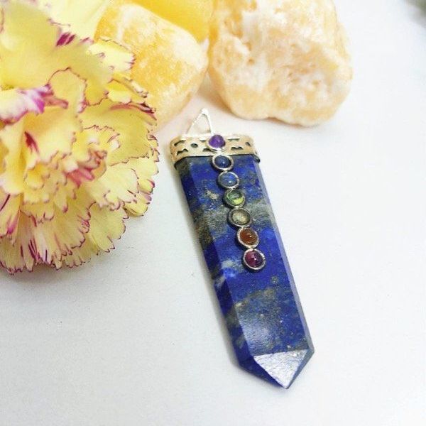 Un colgante de cristal de lapislázuli con piedras de los chakras reposa sobre calcita amarilla. Detrás hay una flor amarilla.