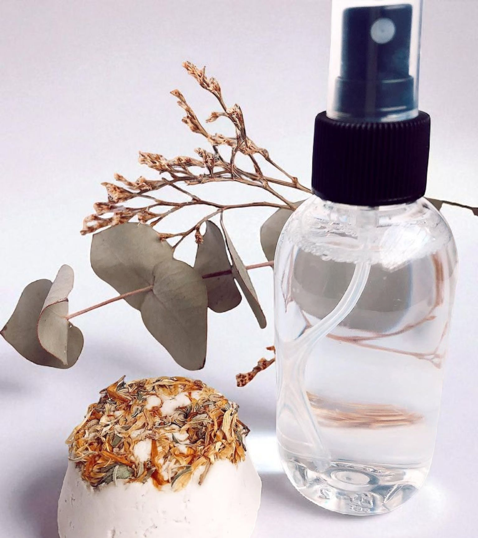 Botella de spray transparente con líquido, ramitas de menta, toalla y frascos sobre una superficie blanca.