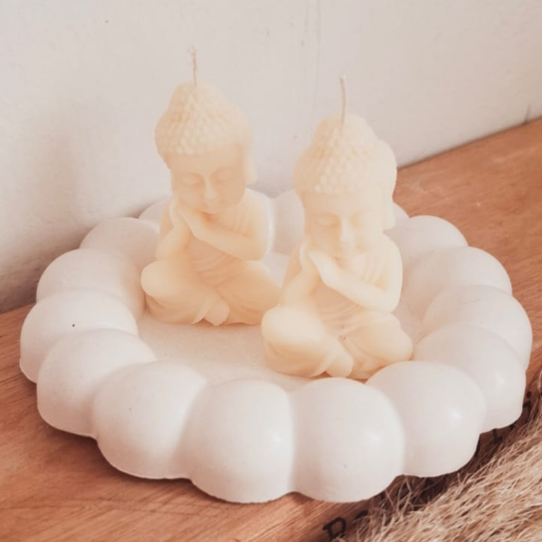 Dos velas esculpidas de color crema sobre un plato blanco con forma de nube, rodeadas de ramitas de lavanda sobre tela morada.