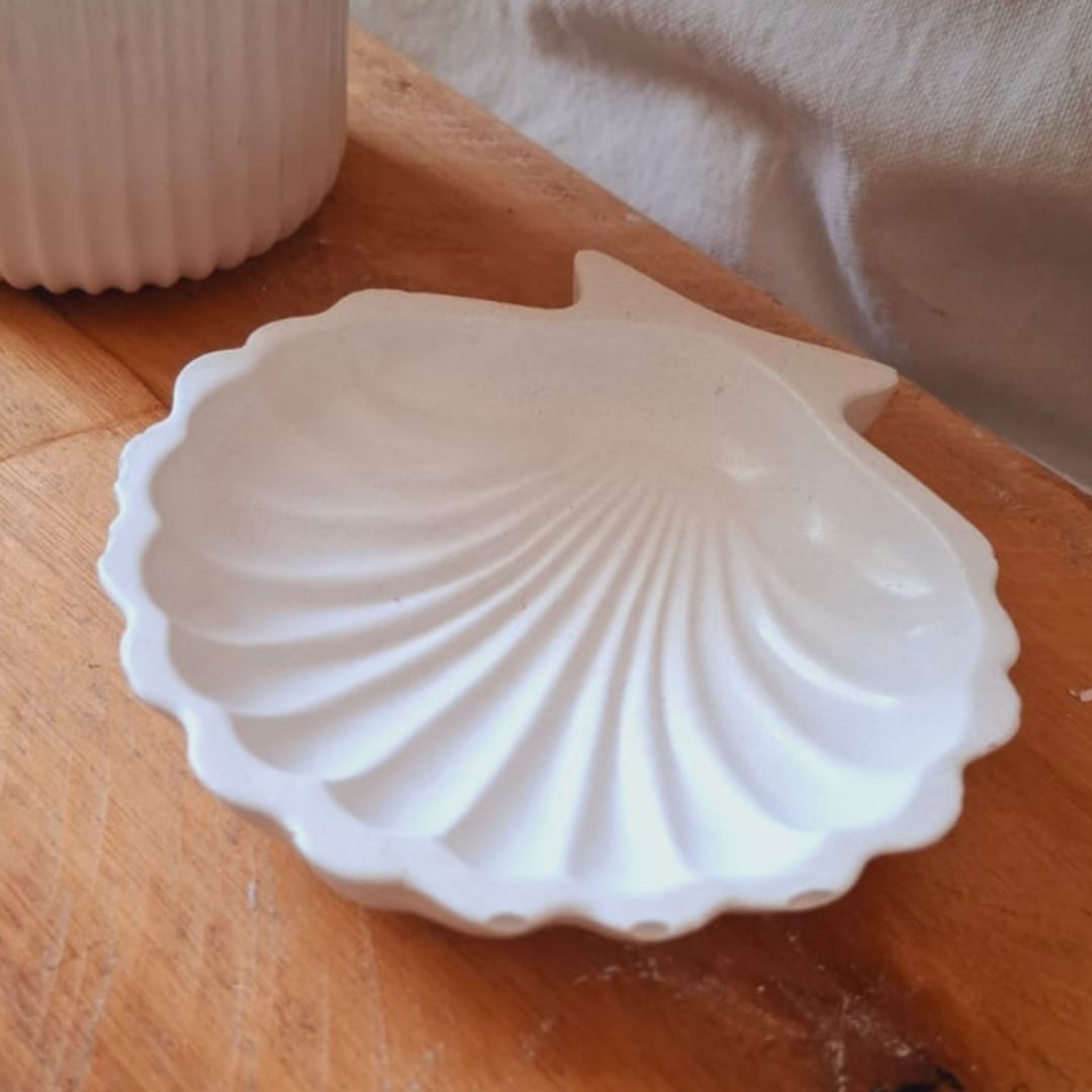 Plato blanco con forma de concha sobre una superficie beige con una vela de burbujas y ramitas de flores blancas al fondo.