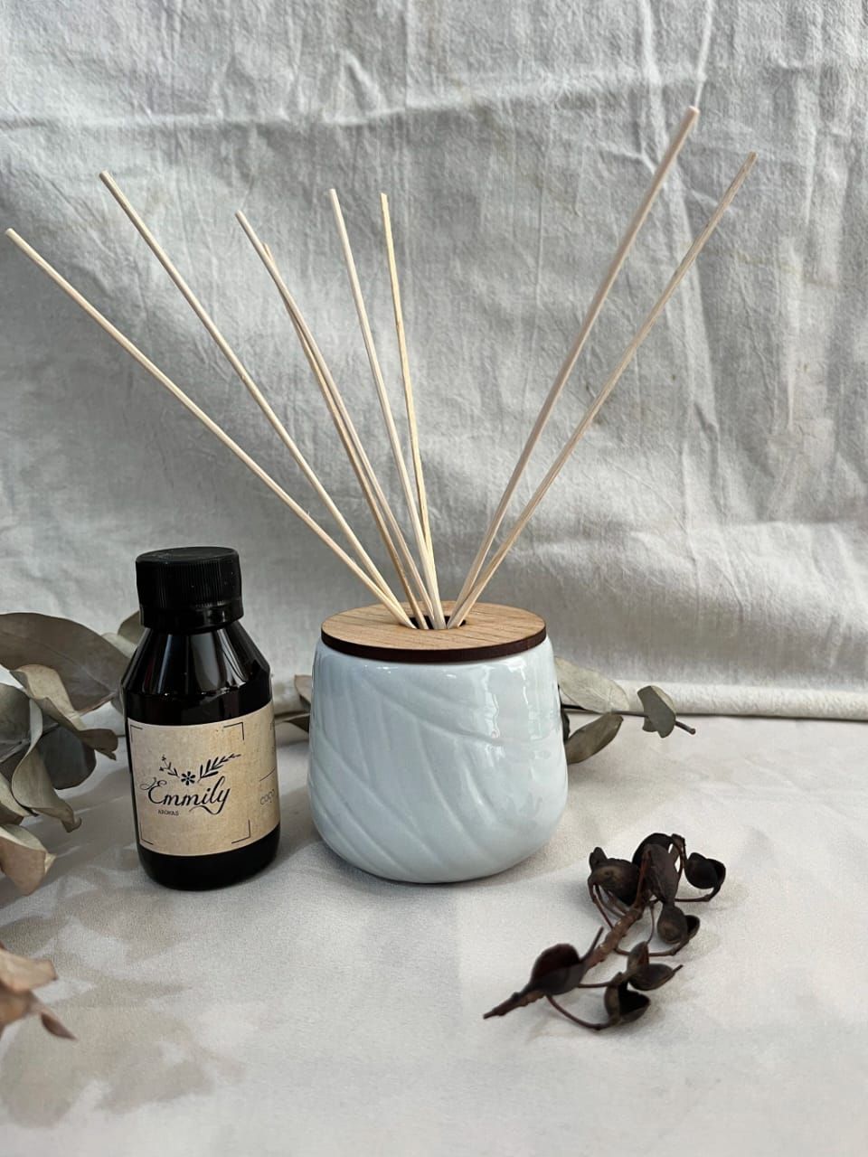 Difusor de varillas con varillas, pequeño frasco de aceite esencial y flores moradas sobre un paño blanco.