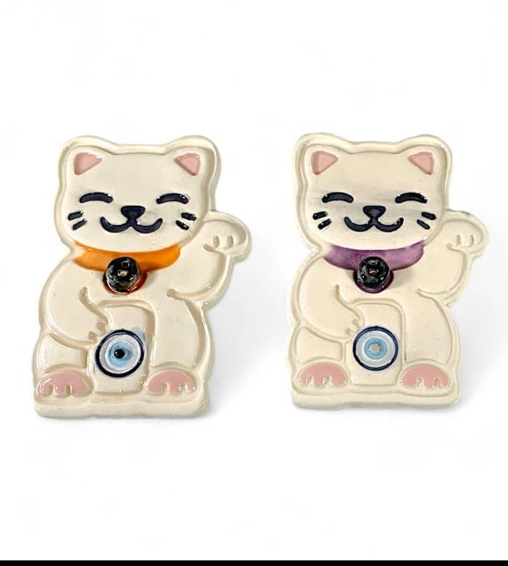 Dos pendientes de Maneki-neko (gato de la suerte) color marfil, uno con collar morado y el otro con rojo, ambos con detalle de ojo azul.