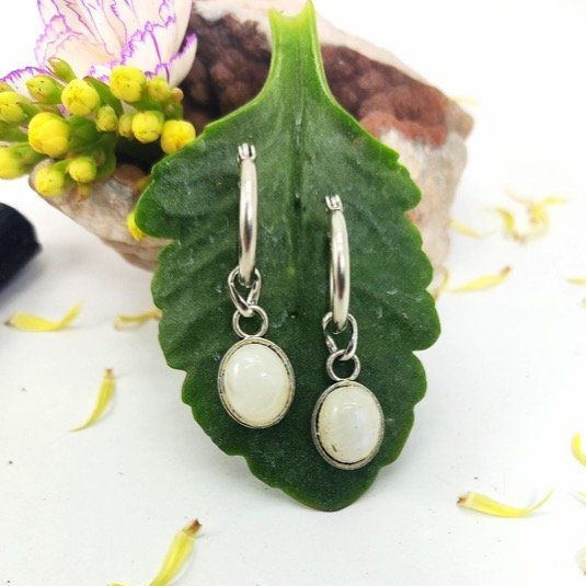 Pendientes de aro plateados con gotas ovaladas de piedra blanca, exhibidos con flores sobre madera flotante.