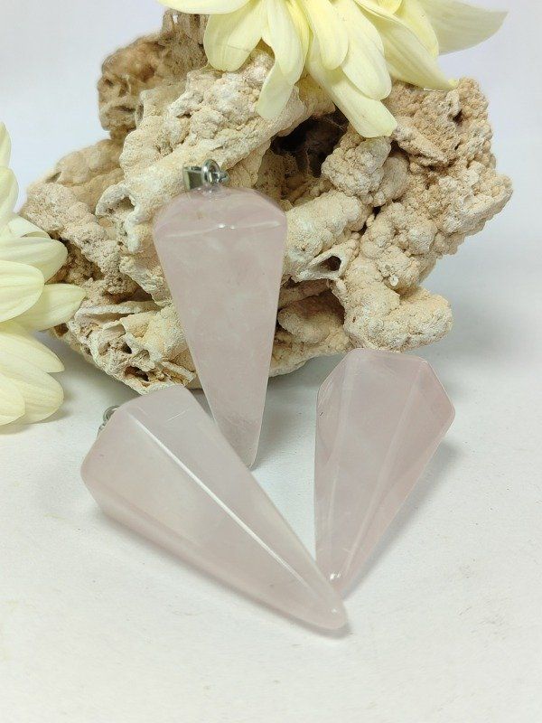 Péndulo de cuarzo rosa con asa de plata, apoyado sobre una roca texturizada de color beige. Una suave flor amarilla al fondo.
