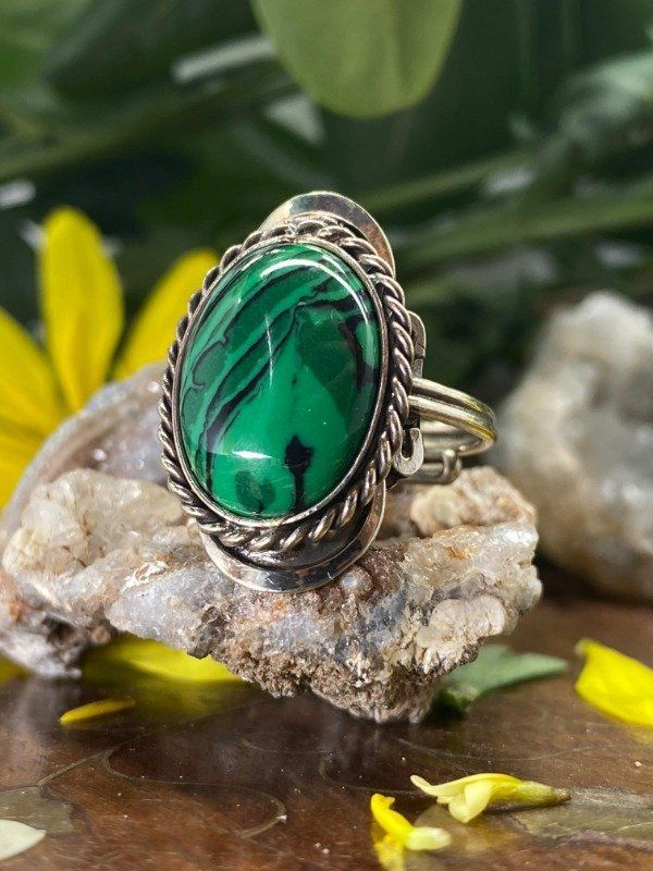 Anillo de piedra malaquita ovalada, engastada en plata, sobre una roca rugosa con fondo floral difuminado.