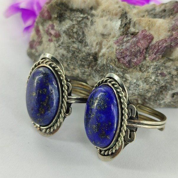 Anillo ovalado de lapislázuli engastado en plata, que descansa sobre una roca rosa y blanca con una flor púrpura difuminada al fondo.