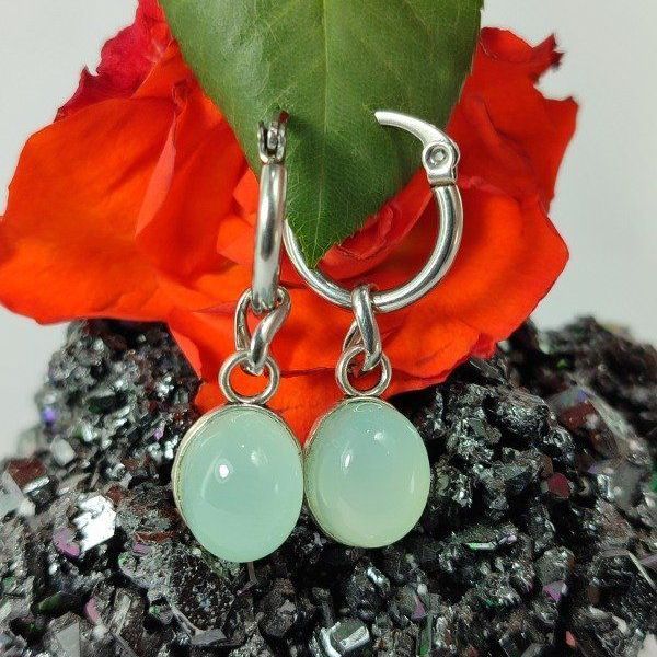 Silver hoop earrings with pale blue-green oval pendants, displayed on a dark, textured surface, next to an orange flower.