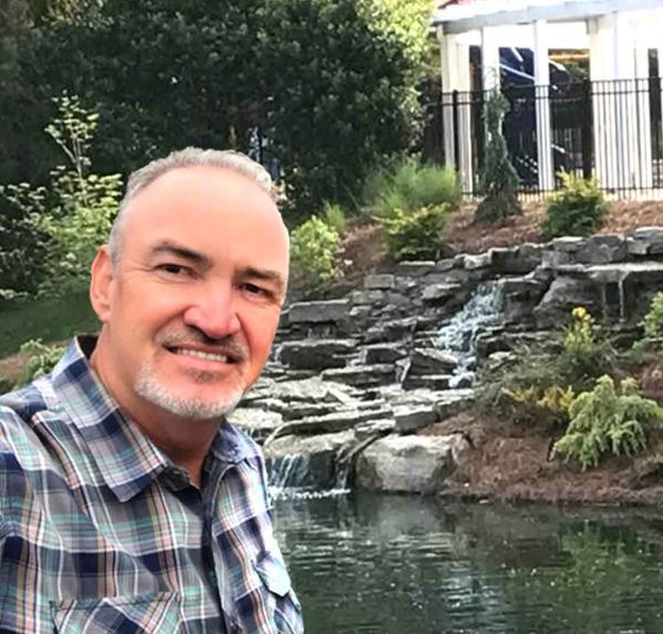 A man in a plaid shirt is taking a selfie in front of a waterfall.