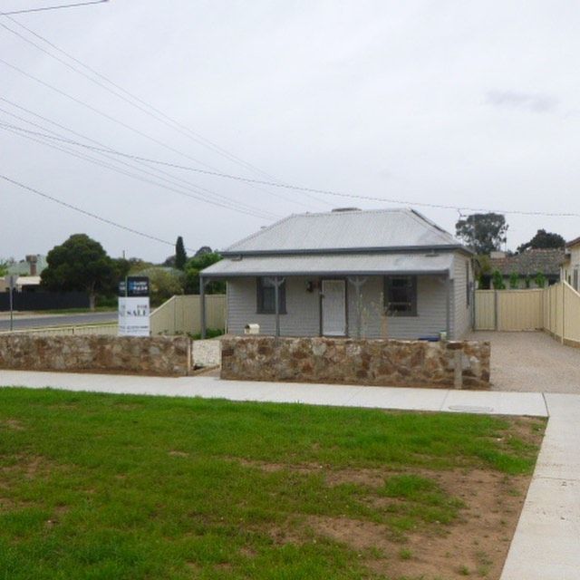 Small gray house with a stone wall in front, a sidewalk, and grass.