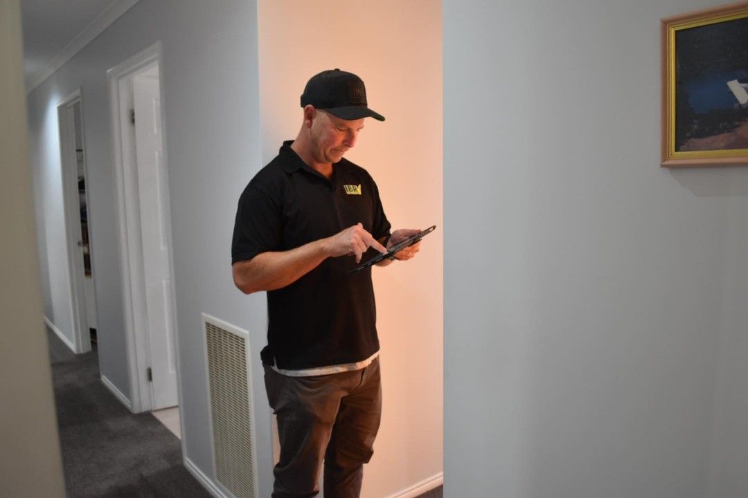 Man in black shirt and cap using a tablet in a hallway with light gray walls and carpet.