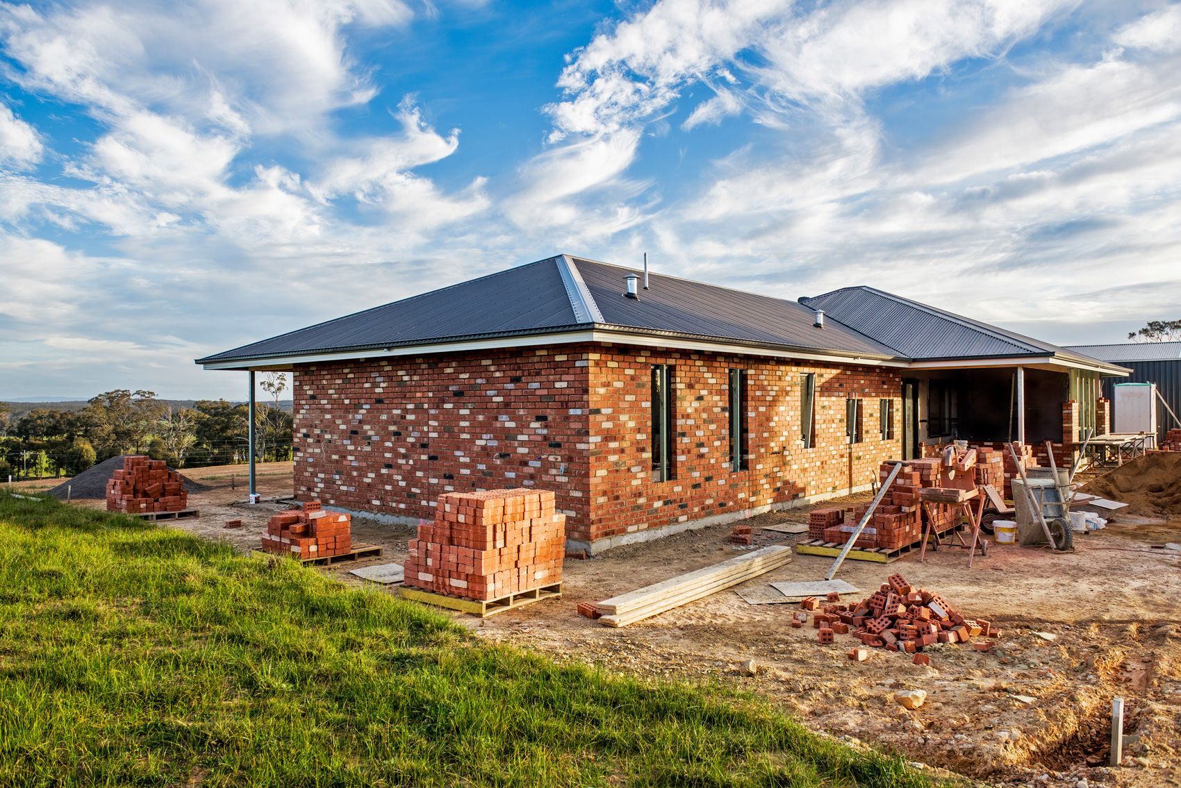 Construction of a brick house with stacked bricks, on a grassy hill, under a partly cloudy sky.