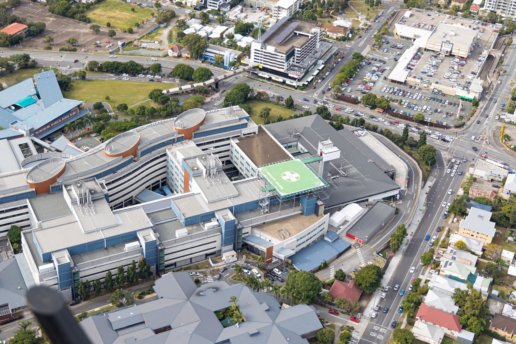 Aerial view of a modern hospital with a helipad, surrounded by buildings, roads, and cars.