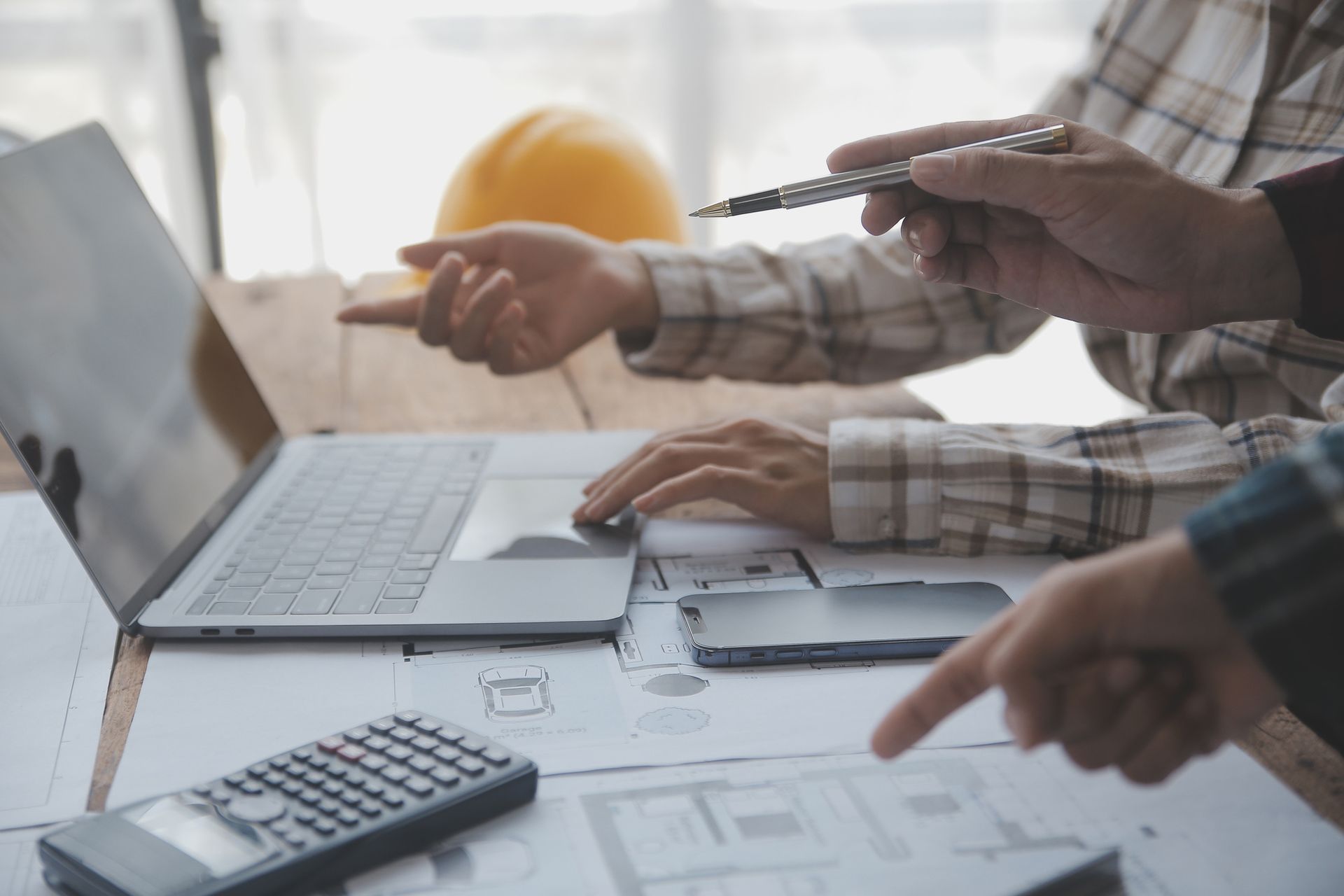 Two people reviewing blueprints and a laptop, pointing, with a calculator and phone on a table.