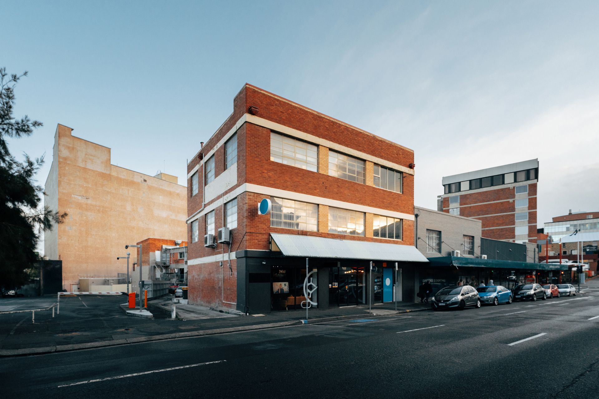 Brick building with blue awning, cars parked on street, brick building to the left and right, overcast sky.
