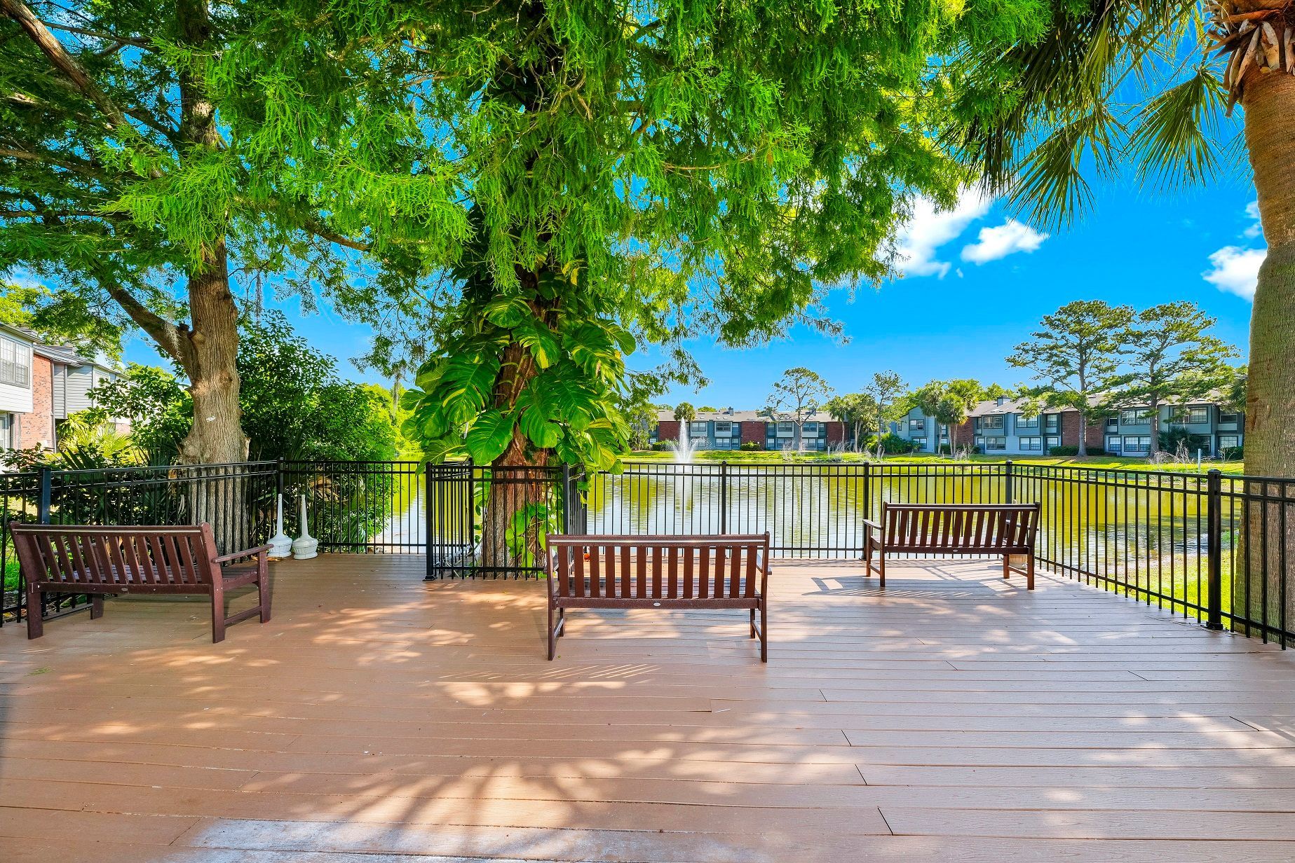 Wooden benches on a deck overlooking a lake with fountain, trees, and buildings under a blue sky at Timberlake in Altamonte Springs, FL.
