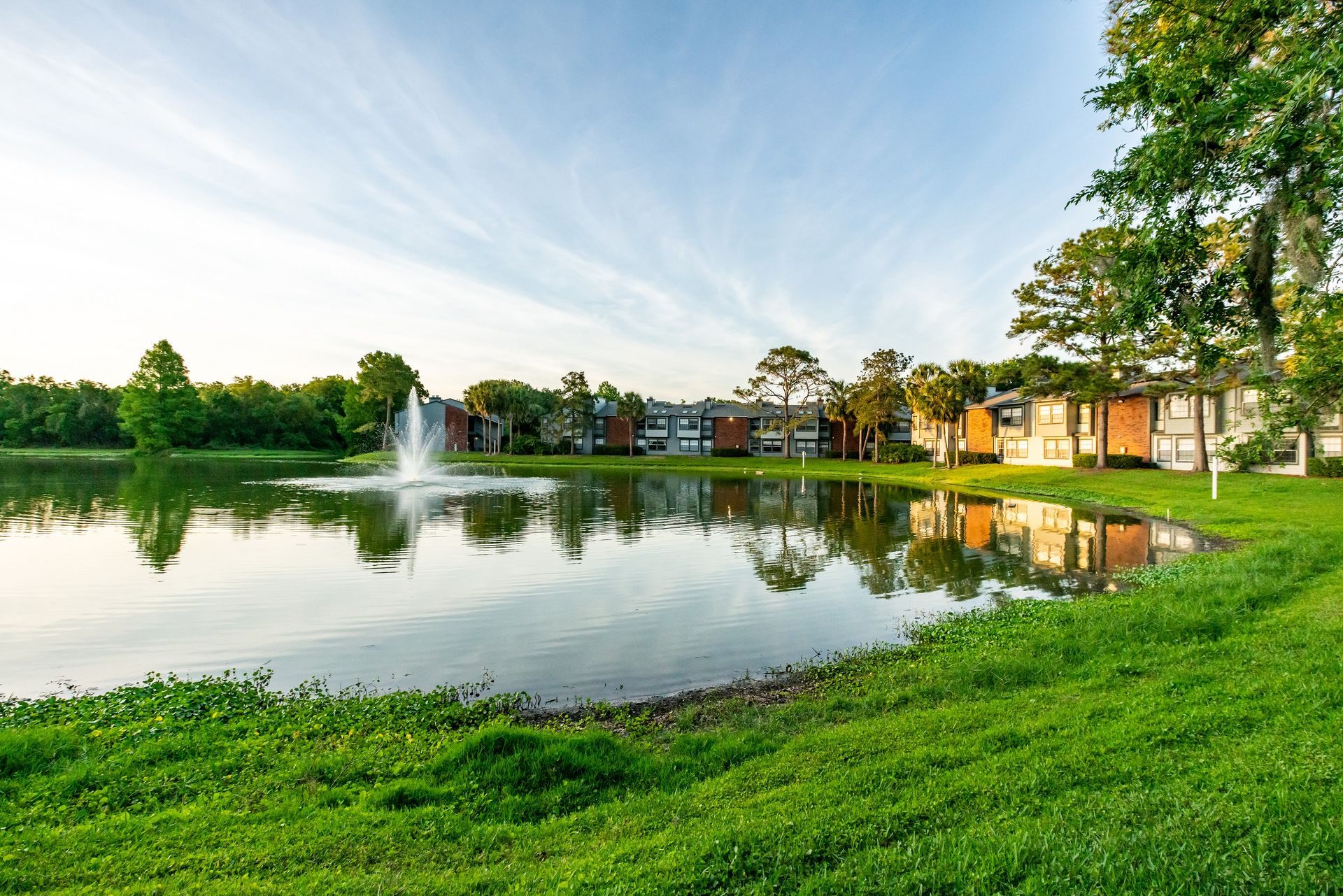 Lakeside apartment complex with a fountain, reflecting buildings, grass, and trees under a blue sky.