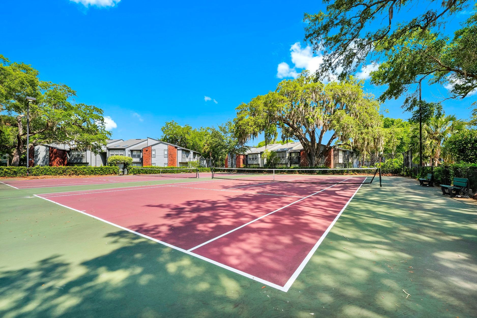 Tennis court with red and green surface, surrounded by trees and buildings under a blue sky at Timberlake in Altamonte Springs, FL.