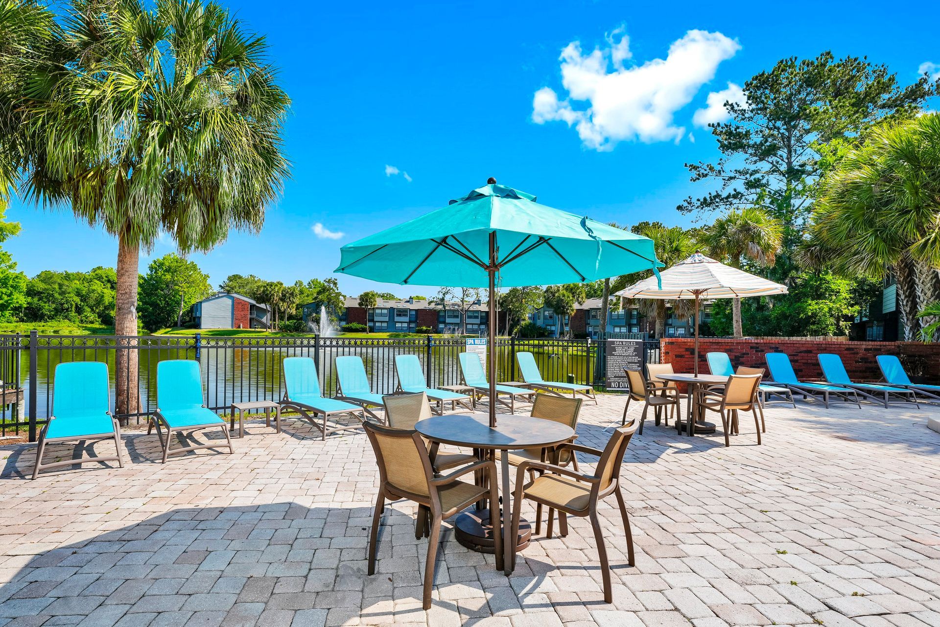 Outdoor seating area by a lake with blue chairs, tables with umbrellas, and palm trees under a sunny sky at Timberlake in Altamonte Springs, FL.