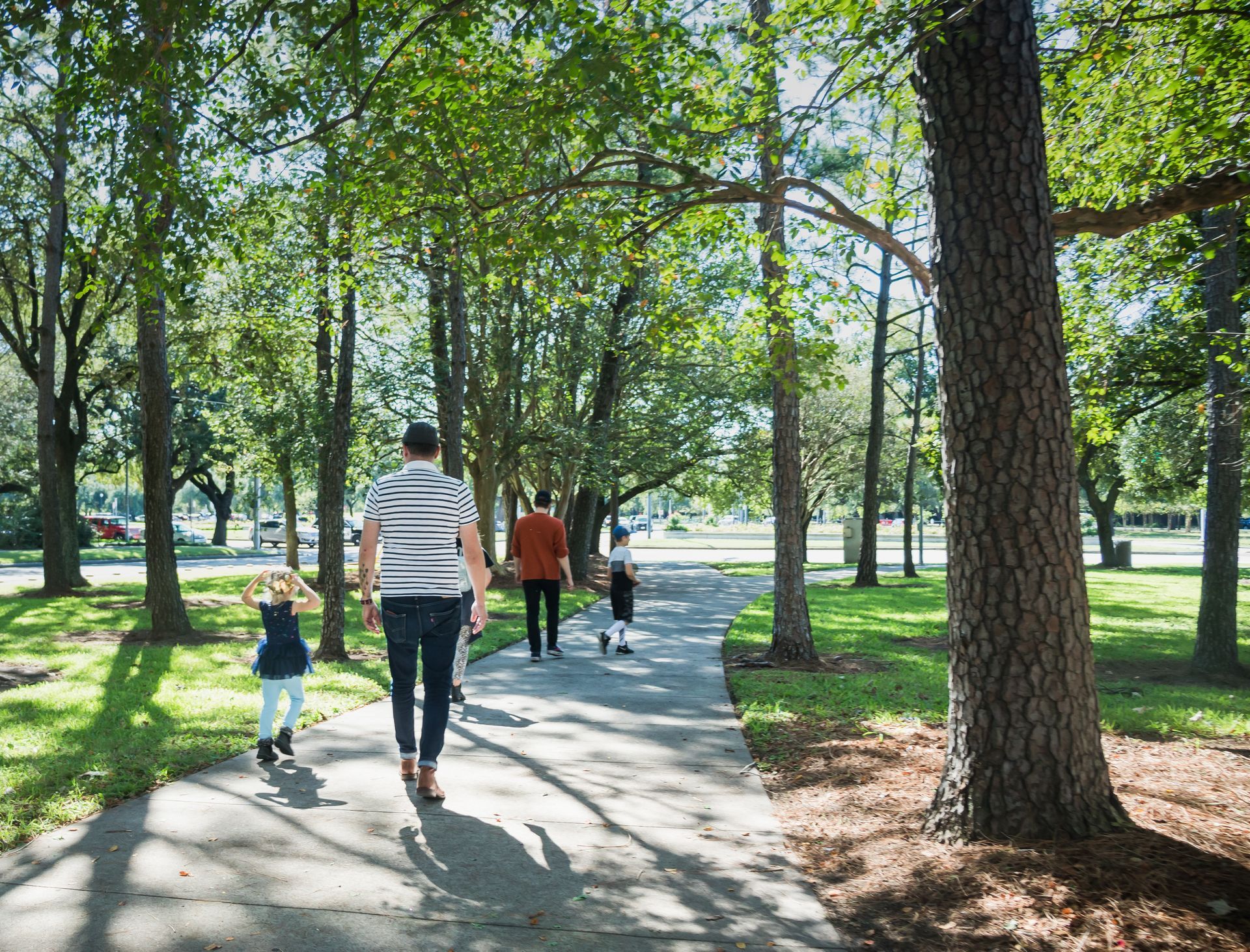 A view from behind of a family walking along a paved pathway winding through a lush, tree-lined park on a sunny day. Large green trees cast dappled shadows across the walkway and the surrounding grassy lawns.
