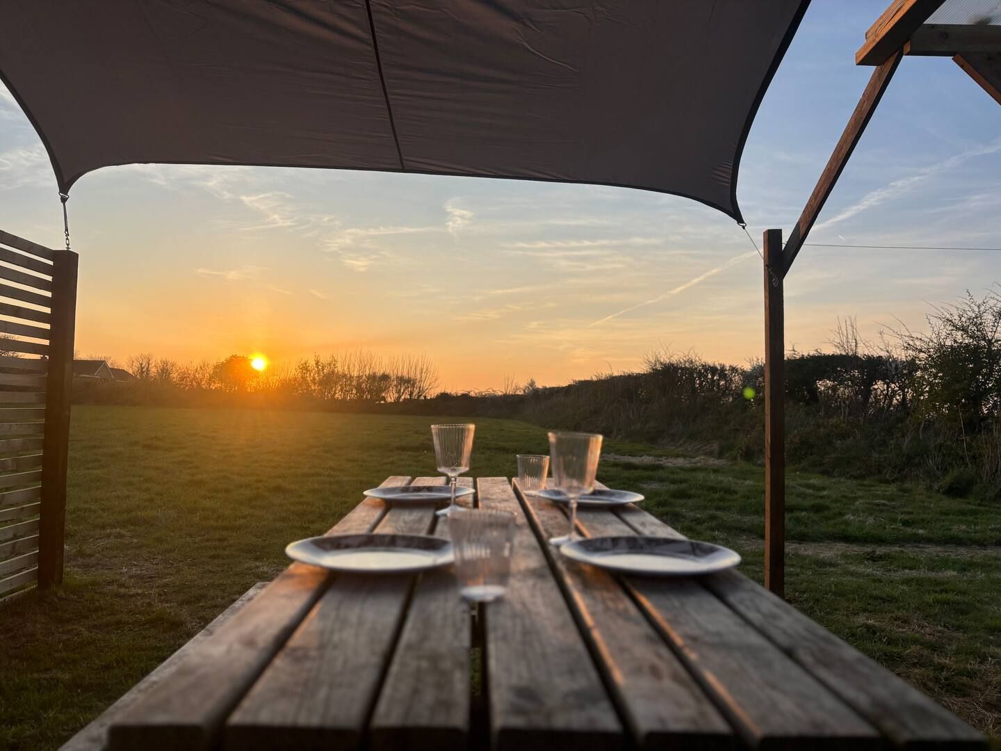 A wooden picnic table with plates and glasses under an umbrella at sunset.