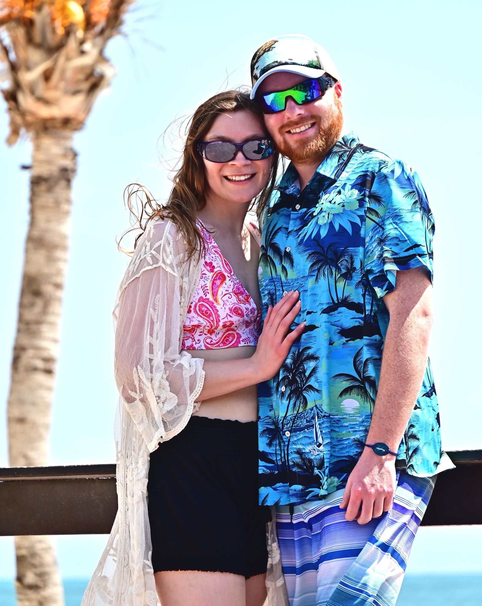 Couple smiles at the camera on a beach. Woman wears sunglasses, cover-up, and shorts. Man in a Hawaiian shirt and sunglasses.