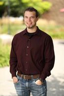 A man in a maroon shirt and blue jeans is standing with his hands in his pockets.