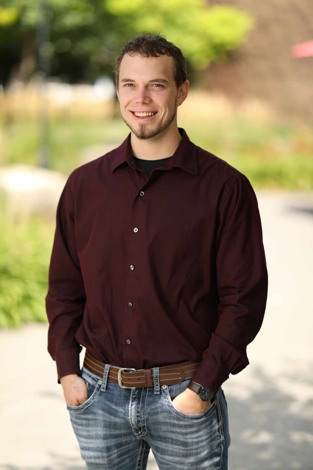 A man in a maroon shirt and blue jeans is standing with his hands in his pockets.