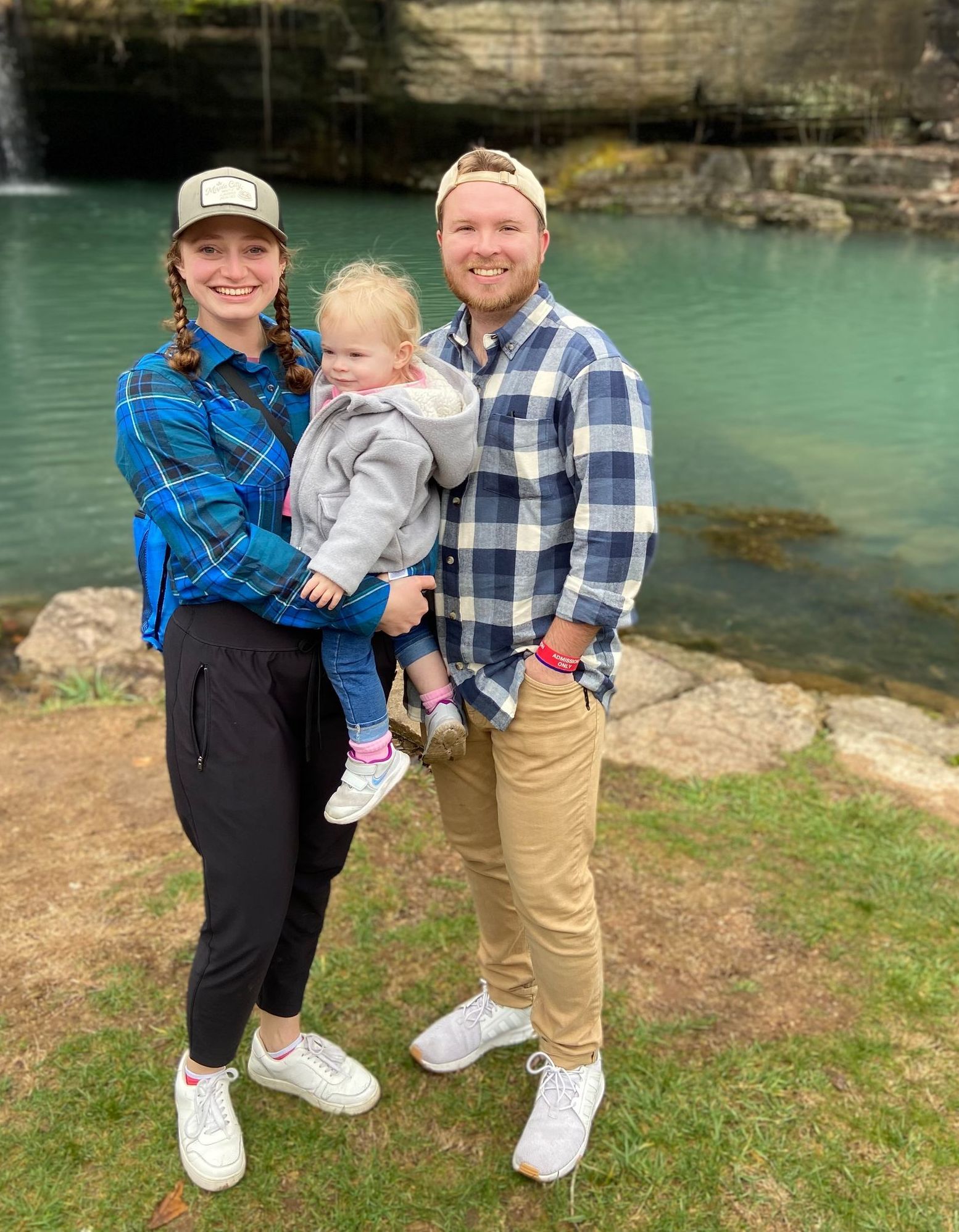 Family of three smiling near a turquoise pond and small waterfall.