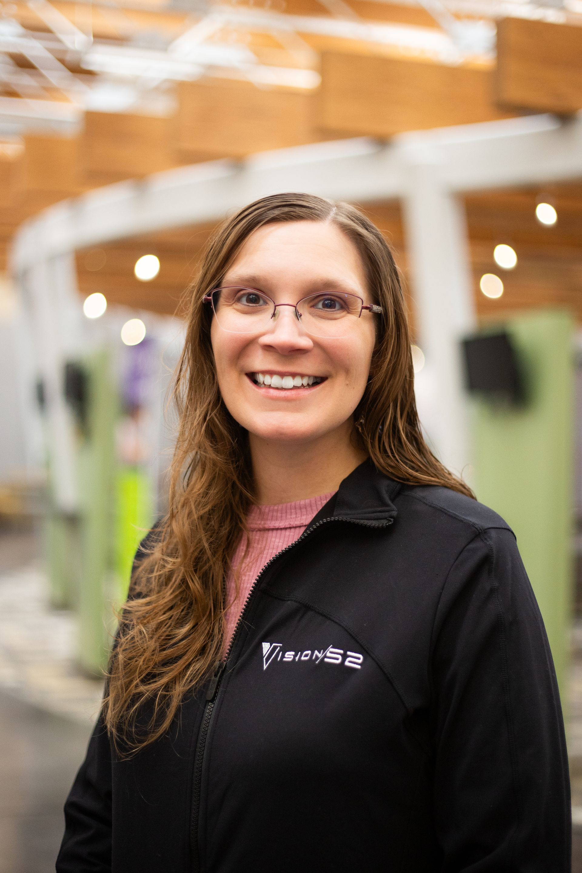 A person with long brown hair and glasses smiles while wearing a black zip-up jacket in a brightly lit indoor space.