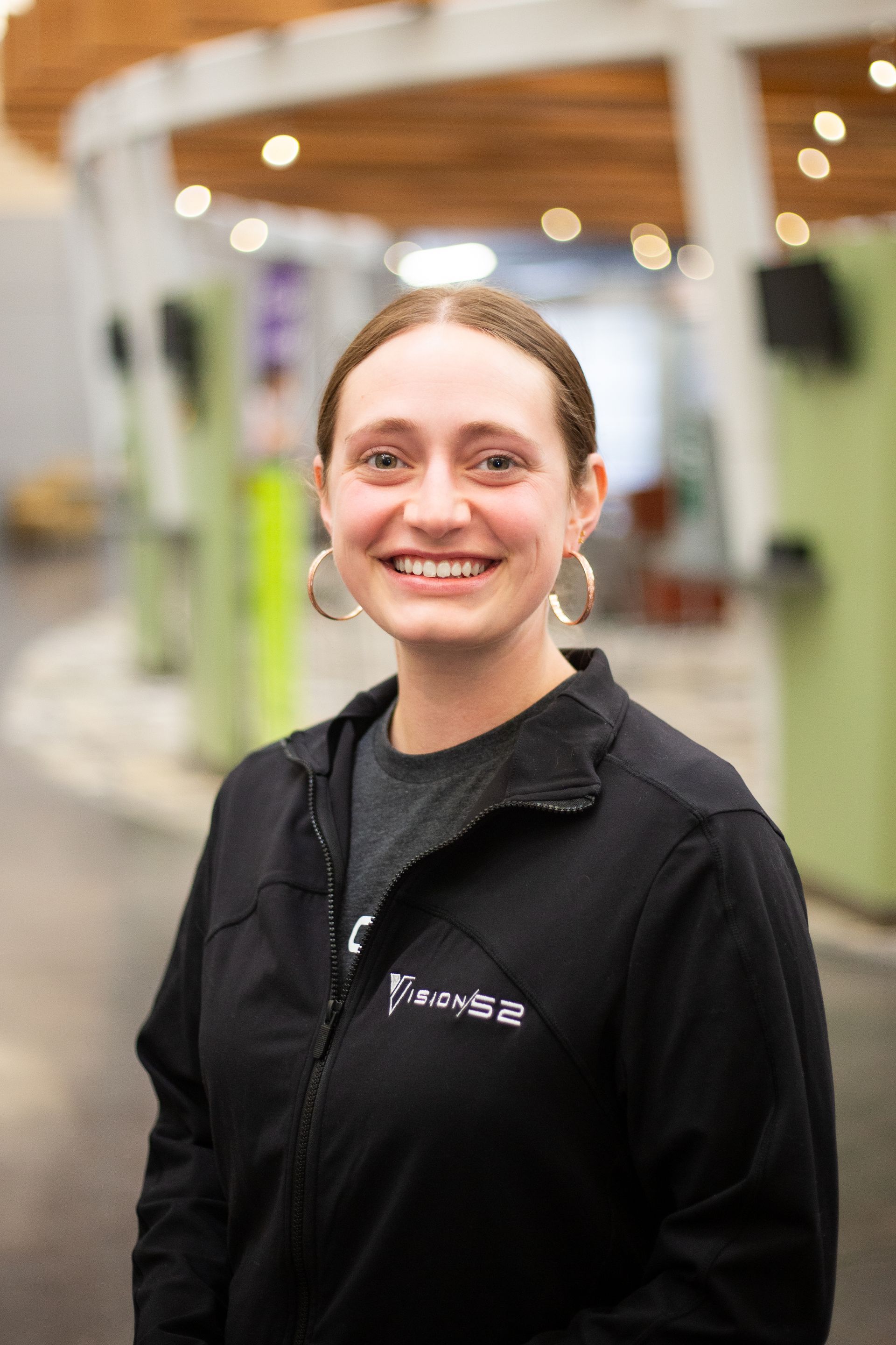 Smiling person with hoop earrings wearing a black zip-up jacket in a brightly lit indoor space.