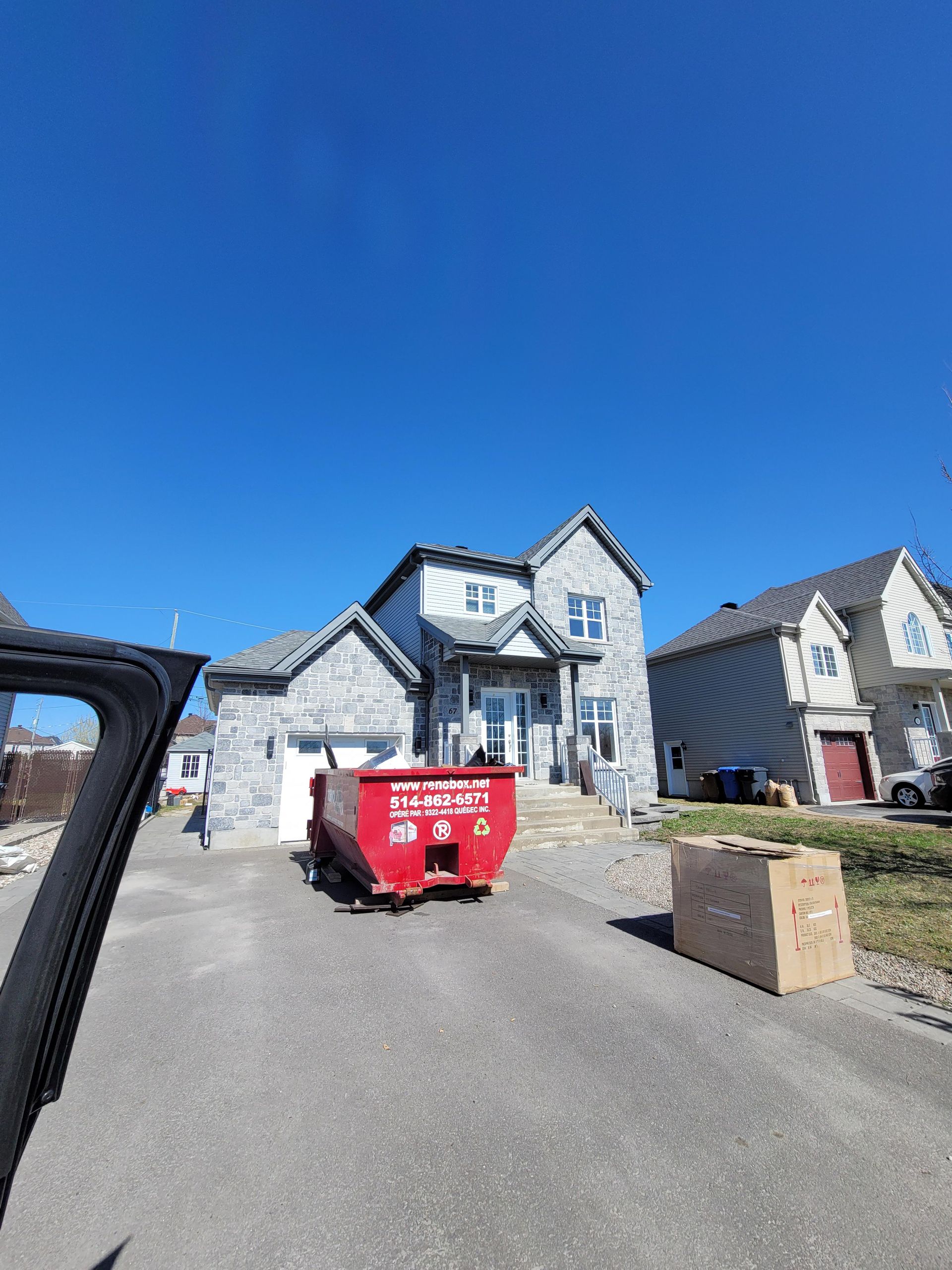 Une maison moderne en pierre sous un ciel bleu azur, avec une benne à ordures rouge dans l'allée et un carton au premier plan.